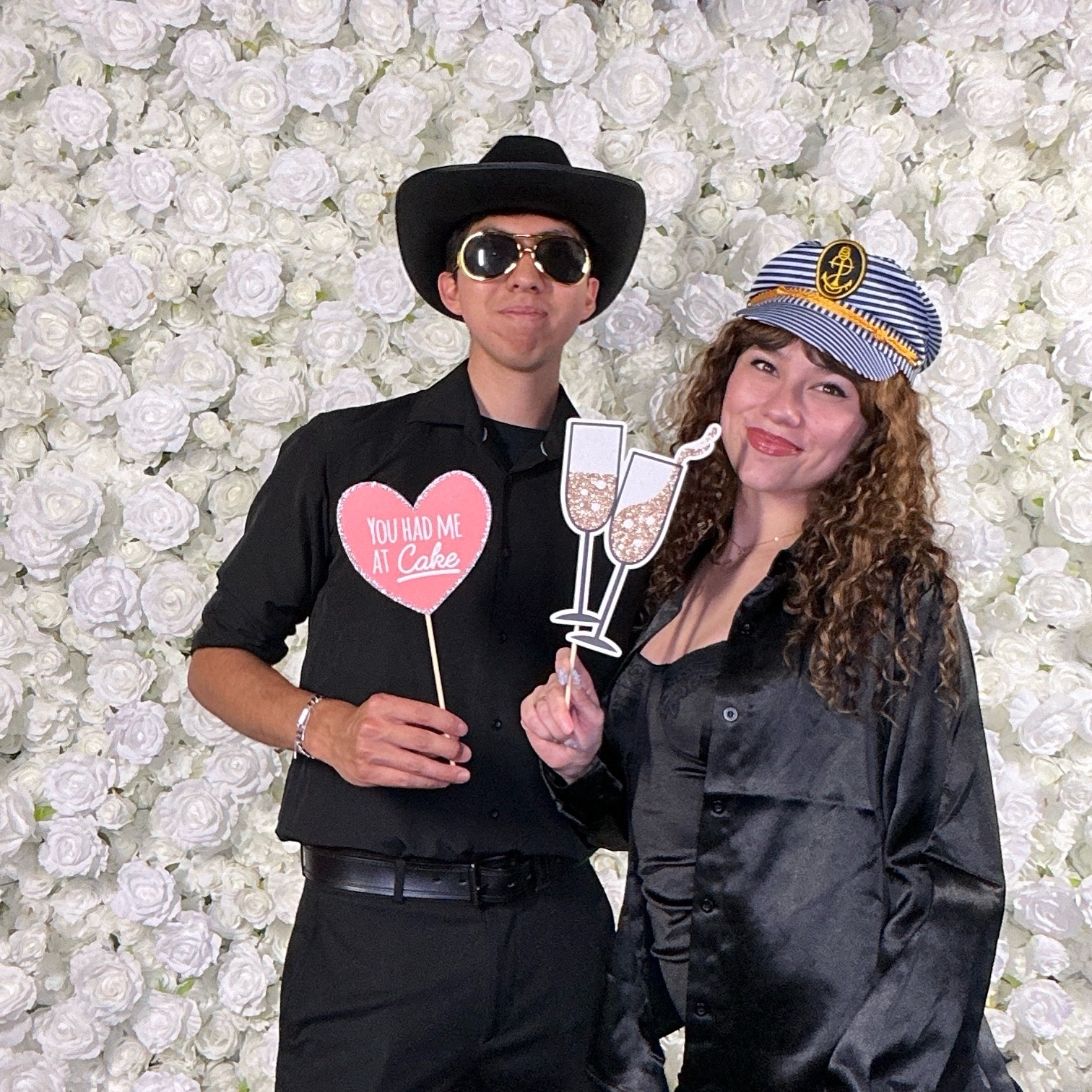 Couple posing with props in front of a white floral wall at an AJM PopBooth